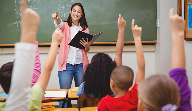 Teacher at front of class, asking students a question, students with hands up