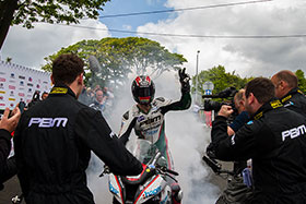 Ian Hutchinson in a cloud of smoke with a helmet on celebrating a TT win with a burnout and one hand in the air while people around film and photograph him