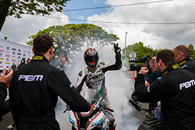 Ian Hutchinson in a cloud of smoke with a helmet on celebrating a TT win with a burnout and one hand in the air while people around film and photograph him
