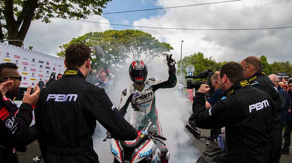 Ian Hutchinson in a cloud of smoke with a helmet on celebrating a TT win with a burnout and one hand in the air while people around film and photograph him