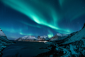 Lake surrounded by snow covered hills, and lights from a nearby town on the far shore. Bright turquoise light streaks through the starry sky.