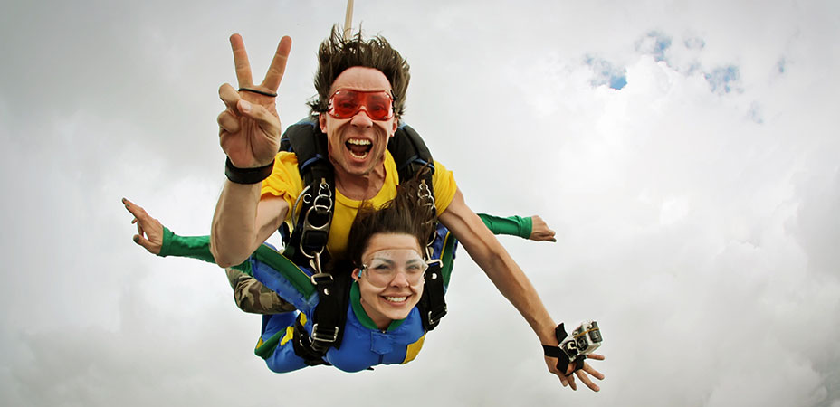 Close up of woman and man skydiving and a cloudy sky. Man is attached to the woman's back and giving peace sign with his hands while the woman is smiling.