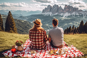 The back of a man and woman sitting on a picnic blanket, surrounded by grass and trees, and looking out over a far reaching mountain range.