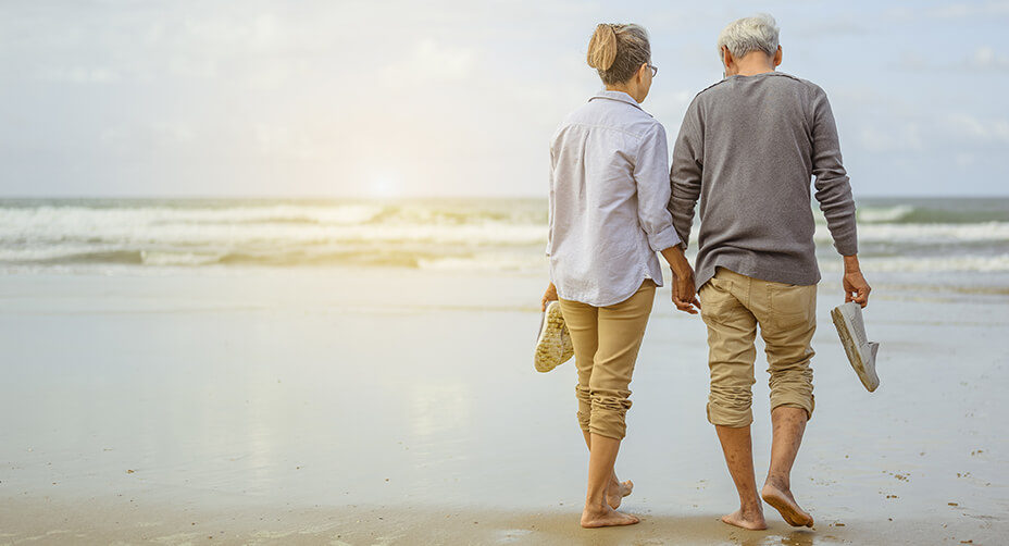 Retired husband and wife walking on beach with shoes in hand